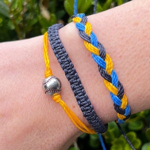 May include: Three friendship bracelets on a wrist. One bracelet is black, one is yellow, and one is blue and yellow braided together. The yellow bracelet has a silver baseball charm.