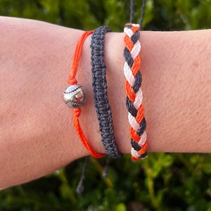 May include: Three friendship bracelets in black, white and orange. One bracelet has a silver baseball charm.
