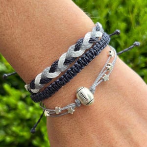 May include: Three bracelets in shades of gray and white. The top bracelet is braided, the middle one is a macrame style, and the bottom one features a football charm and star-shaped beads. The bracelets are on a wrist.