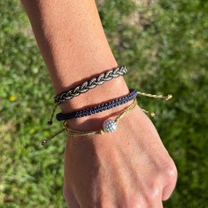Puede incluir: Tres pulseras trenzadas en tonos azul marino y dorado se muestran en una muñeca. Una pulsera presenta una cuenta esférica plateada. Las pulseras están sobre un fondo verde borroso, lo que sugiere un entorno al aire libre. Las pulseras son hechas a mano.