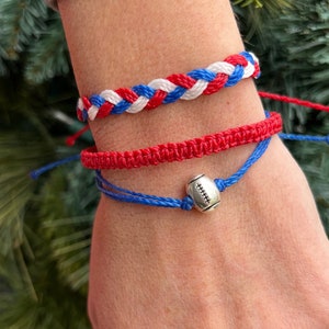 May include: Three friendship bracelets in red, white, and blue colors. The top bracelet is a braided design. The middle bracelet is a solid red color. The bottom bracelet is blue with a silver football charm.