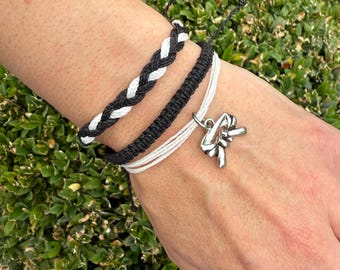 May include: A collection of handmade bracelets in black and white. One bracelet is braided, another is a solid black cord, and a third is white. A silver charm hangs from the white bracelet. The bracelets are displayed on a person's wrist.