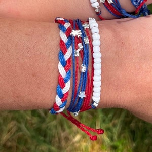 Puede incluir: Un conjunto de pulseras de amistad coloridas con cordones trenzados rojo, blanco y azul y una pulsera de cuentas blancas con abalorios de estrellas plateadas.