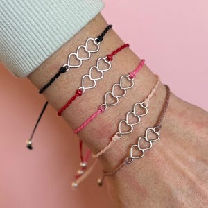May include: Four friendship bracelets with silver heart charms on a pink background. The bracelets are black, red, pink, and tan, with adjustable ties. The charms feature three connected hearts.