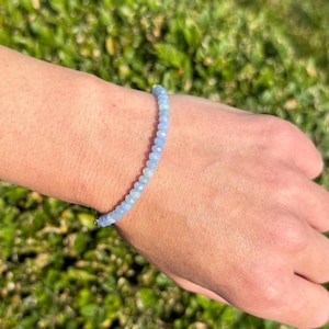 May include: A delicate bracelet composed of small, light blue beads. The bracelet is worn on a wrist, with a green, blurred background. The beads appear to be faceted, catching the light. The bracelet is a simple, elegant piece of jewelry.