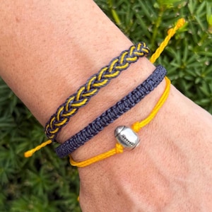Puede incluir: Tres pulseras en azul marino y amarillo. La pulsera superior está trenzada con hilos amarillos y azul marino. La pulsera del medio es azul marino. La pulsera inferior es amarilla con un dije de fútbol plateado. Las pulseras están en una muñeca.