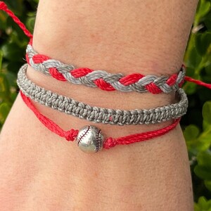 May include: Three friendship bracelets on a wrist. The top bracelet is gray and red braided together. The middle bracelet is gray and the bottom bracelet is red with a silver baseball charm.