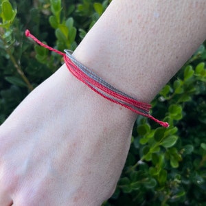 May include: A red and gray friendship bracelet on a person's wrist.