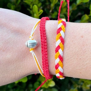 Könnte beinhalten: Zwei Freundschaftsarmbänder, eines rot und eines mit roten, weißen und gelben geflochtenen Strängen. Ein silberner Anhänger in Form eines Fußballs ist an dem roten Armband befestigt.