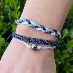 May include: Two friendship bracelets with a baseball charm. The top bracelet is black and white braided, and the bottom bracelet is black with a white string. The charm is silver with a baseball design.