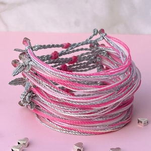 May include: A stack of pink, white, and silver friendship bracelets with silver feather charms. The bracelets are on a pink surface with silver heart beads scattered around them.