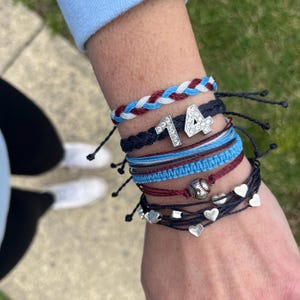 May include: A stack of colourful friendship bracelets with a silver charm that says "14" with rhinestones. The bracelets are made of braided string and include a baseball charm.