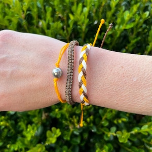 Könnte beinhalten: Drei Freundschaftsarmbänder in den Farben Braun, Weiß und Gelb. Die Armbänder sind geflochten und haben einen silbernen Perlenanhänger.