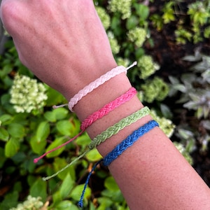 Peut inclure: Quatre bracelets d'amitié tressés de couleur rose, rouge, vert et bleu sur le poignet d'une personne. Les bracelets sont attachés ensemble aux extrémités.