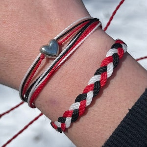 Puede incluir: Se muestran dos pulseras. Una pulsera tiene un dije de corazón plateado y está hecha de cordones rojos, negros y blancos. La otra pulsera está trenzada con cordones rojos, negros y blancos. Las pulseras están en una muñeca.