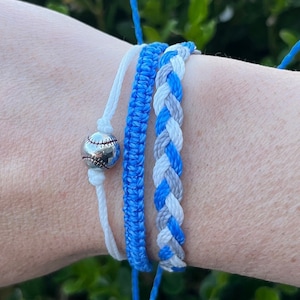 May include: Three friendship bracelets in blue, white, and gray colors. The bracelets are braided and tied together with a silver baseball charm.