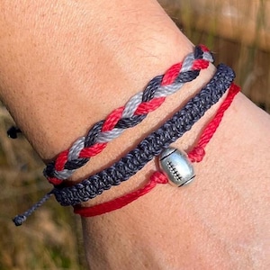 May include: Three braided friendship bracelets in red, gray, and black with a silver football charm. The bracelets are worn on a person's wrist.