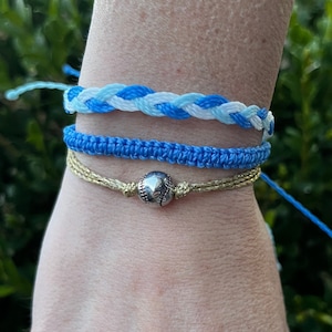 May include: Three friendship bracelets on a wrist. The top bracelet is blue, white, and light blue braided together. The middle bracelet is a solid blue. The bottom bracelet is a gold cord with a silver baseball charm.