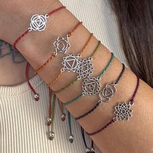 Op de afbeelding: Een collectie van zeven chakra armbanden. Elke armband heeft een andere kleur koord en een zilverkleurige bedel met een uniek chakra symbool. De armbanden zijn verstelbaar en vastgemaakt met een schuifknoop.