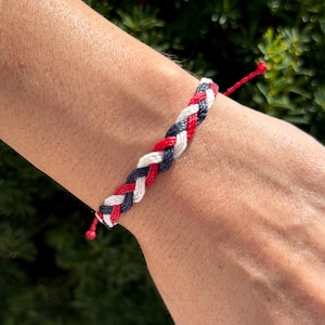 May include: Braided bracelet featuring red, white, and navy blue threads. The bracelet is hand-crafted with a woven design, and the ends are finished with red knots. This accessory is suitable for everyday wear.