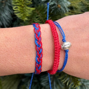 Könnte beinhalten: Drei handgefertigte Armbänder in Rot und Blau. Eines ist mit roten und blauen Fäden geflochten. Ein weiteres ist ein rotes Makramee-Armband. Das dritte ist blau mit einem silbernen Football-Charm. Die Armbänder werden an einem Handgelenk getragen.