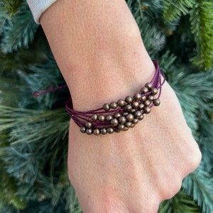 May include: A handmade bracelet featuring multiple strands of deep purple cord. The bracelet is adorned with a cluster of small, round, bronze-colored beads. The bracelet is worn on a wrist, showcasing the design and color contrast.