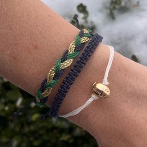 Pode incluir: Três pulseiras de amizade em um pulso. Uma pulseira é branca com um amuleto de futebol dourado. As outras duas pulseiras são trançadas com fios verdes, dourados e azul marinho.