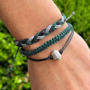 May include: Three layered bracelets with black, gray, and green string. The bottom bracelet has a silver football charm.