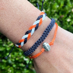 May include: Three friendship bracelets in orange, white, and navy blue colors. The bracelets are braided and knotted. One bracelet has a silver football charm.