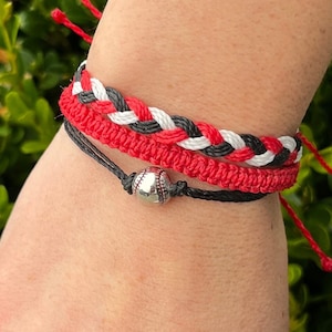 Puede incluir: Un brazalete de amistad trenzado rojo, blanco y negro con un brazalete de cuerda negro con un dije de béisbol plateado.