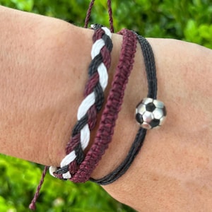 Puede incluir: Tres pulseras de amistad trenzadas en granate, negro y blanco con un dije de balón de fútbol plateado.