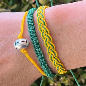 Może przedstawiać: Trzy bransoletki przyjaźni w kolorze zielonym i żółtym z srebrnym zawieszką w kształcie piłki nożnej. Bransoletki są wykonane z plecionego sznurka.
