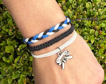 May include: A collection of braided bracelets in black, white, and blue. One bracelet features a silver charm. The bracelets are displayed on a wrist against a backdrop of green foliage. The bracelets are handmade.