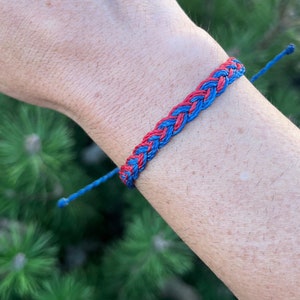 May include: Braided bracelet featuring a vibrant pattern of interwoven red and blue threads. The bracelet has adjustable blue cords on each end for a customizable fit. The design is simple and eye-catching, suitable for everyday wear.