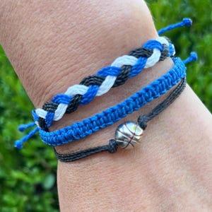 Könnte beinhalten: Drei geflochtene Freundschaftsarmbänder in Blau, Weiß und Schwarz mit einem silbernen Basketball-Anhänger. Die Armbänder sind auf dem Handgelenk einer Person übereinander gelegt.