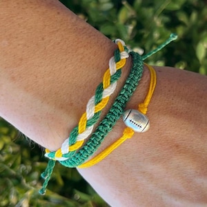 May include: Three friendship bracelets in green, white, and yellow with a silver football charm. The bracelets are tied on a person's wrist.