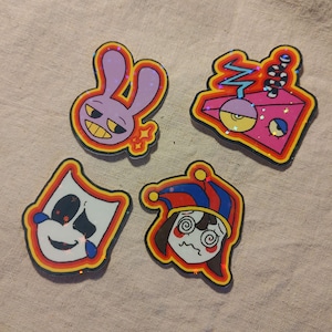 May include: Four colorful, glittery cartoon character pins. The pins include a purple bunny, a pink and yellow geometric shape with a snake, a white mask with black accents, and a jester-like character with a red, blue, and yellow hat.