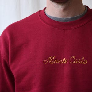 May include: A burgundy crew neck sweatshirt with the embroidered text "Monte Carlo" in gold script. The sweatshirt has a ribbed neckline and a relaxed fit. The model is wearing a gray undershirt.