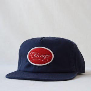 May include: Navy blue baseball cap with a flat brim. The cap features a red oval patch with the word "Chicago" embroidered in white script. The hat is a classic style, suitable for casual wear.