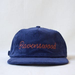 May include: A navy blue corduroy baseball cap with the word "Ravenswood" embroidered in orange script across the front. The cap has a curved brim and a small adjustable strap at the back.