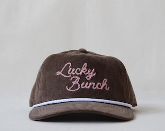 May include: A brown corduroy baseball cap featuring the text Lucky Bunch embroidered in pink script across the front panel. The cap includes a white braided rope detail running horizontally across the base of the crown above the brim.
