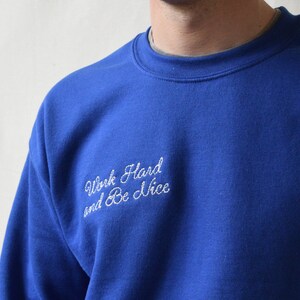 May include: A royal blue sweatshirt with the embroidered text "Work Hard and Be Nice" in white script. The crew neck sweatshirt is a solid color and appears to be made of a soft material.