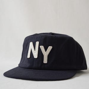 May include: Navy blue baseball cap with the letters "NY" in white. The hat has a curved brim and a classic, casual style. The letters are stitched onto the front of the cap.