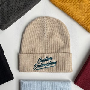 May include: A beige ribbed beanie with the text "Custom Embroidery" in green cursive lettering.