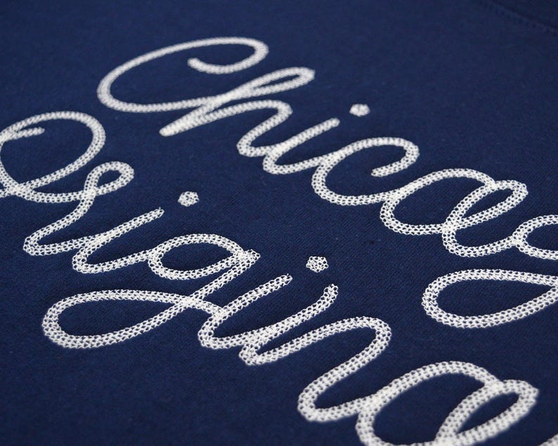 Puede incluir: Primer plano de una tela azul marino con las palabras "Chicago Originals" bordadas en blanco con letra cursiva. La costura tiene un dise&ntilde;o calado delicado, creando un efecto texturizado sobre el fondo oscuro.