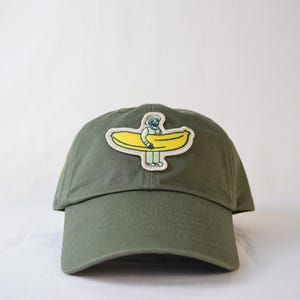 May include: Olive green baseball cap with a patch featuring a cartoon figure carrying a yellow banana. The patch has a white border and the figure is wearing a hat and green pants. The cap is made of a soft material.