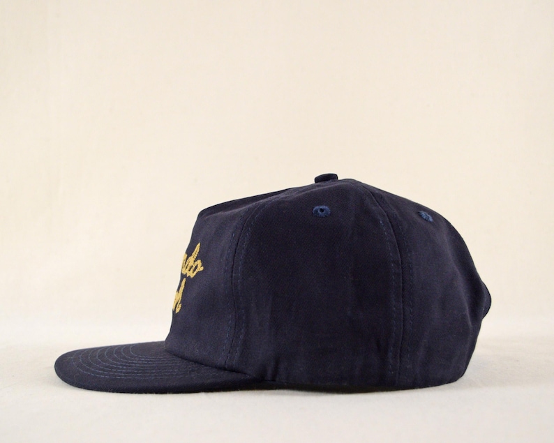 Puede incluir: Gorra de b&eacute;isbol azul marino con visera plana. La gorra presenta texto dorado bordado en la parte delantera. La gorra tiene un bot&oacute;n en la parte superior y una visera curva. Es un estilo cl&aacute;sico.
