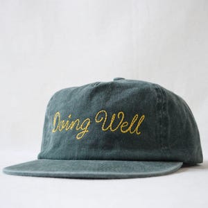 May include: A teal-colored, unstructured baseball cap with a flat brim. The phrase "Doing Well" is embroidered in a yellow, cursive font across the front of the cap. The cap is made of a soft, textured fabric.