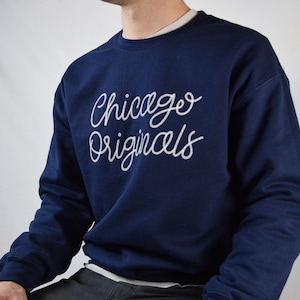 Puede incluir: Sudadera azul marino con la inscripci&oacute;n "Chicago Originals" en blanco en el pecho. La sudadera tiene cuello redondo y mangas largas. La persona lleva una camiseta interior blanca.
