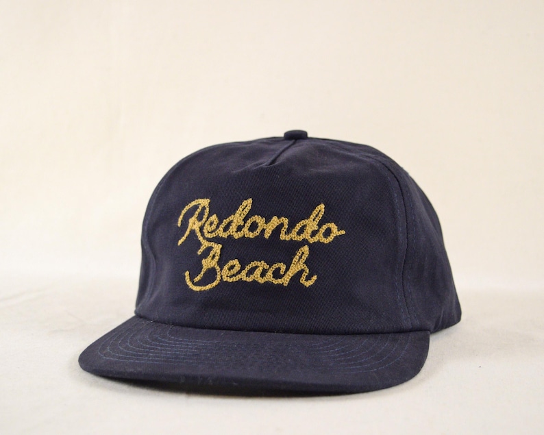 Puede incluir: Gorra de b&eacute;isbol azul marino con visera curva. La gorra presenta el texto bordado "Redondo Beach" en oro en la parte delantera. La gorra tiene un bot&oacute;n en la parte superior y un ajuste estructurado.